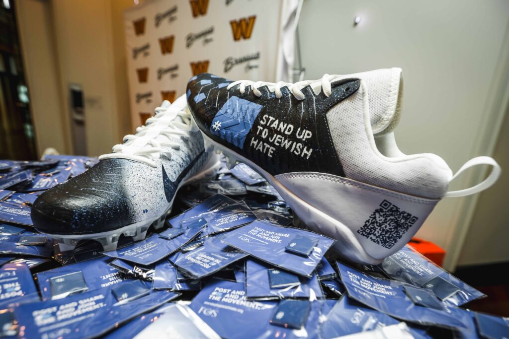 A pair of Nike football cleats featuring the words “Stand Up to Jewish Hate” displayed on one shoe, surrounded by campaign cards. The cleats are black, blue, and white with a QR code printed on the heel.